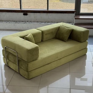 May include: A modern, olive green sectional sofa with a corduroy texture. The sofa has a modular design with a low profile and a matching square pillow. A silver metal frame supports the armrest.