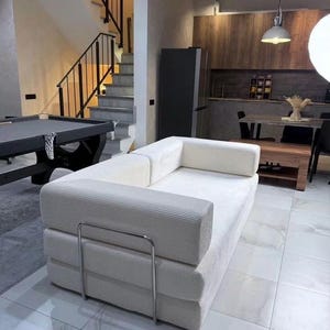 Può includere: Un moderno soggiorno con un divano in velluto a coste color crema con struttura in acciaio inossidabile. La stanza comprende un tavolo da biliardo, una scala e una zona cucina con mobili in legno e un tavolo da pranzo. Il pavimento è piastrellato.