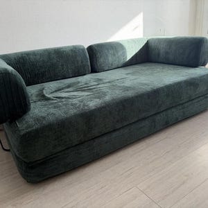 以下が含まれることがあります： 丸みを帯びたアームレストが付いた、濃いティール色のコーデュロイの長椅子。ソファはロープロファイルでシンプルなデザインです。フレームは細い黒い金属製の脚で支えられています。長椅子は、明るい色の床と白い壁のある部屋に置かれています。