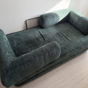 以下が含まれることがあります： 丸みを帯びたアームレストが付いた濃いティール色のラウンジチェア。 張り地は質感があり、白い壁と明るい色の床に設置されています。 デザインはローバックで、快適で魅力的な美学が特徴です。