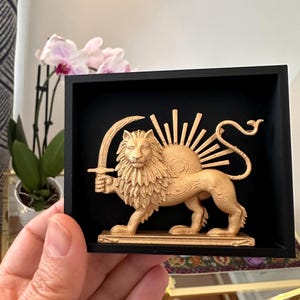 May include: A gold-coloured lion sculpture holding a sword, set against a black background within a square frame. The lion has a detailed mane and a sun-like design behind its head. The sculpture is held by a hand.