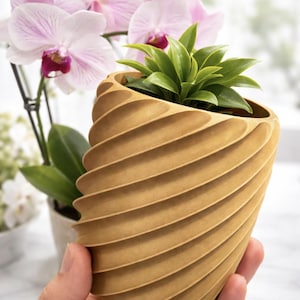Peut inclure: Un pot de fleur marron clair en spirale contenant une petite plante verte. Le pot a un motif à rainures et est tenu devant un arrière-plan flou avec des orchidées roses et d'autres plantes. Le pot mesure environ 13 cm de haut.