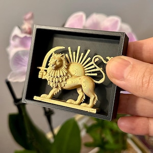 May include: A gold-coloured lion and sun emblem, set against a black background within a square frame. The lion holds a sword, and the sun has radiating beams. The frame is held by a hand, with a blurred background of plants and flowers.