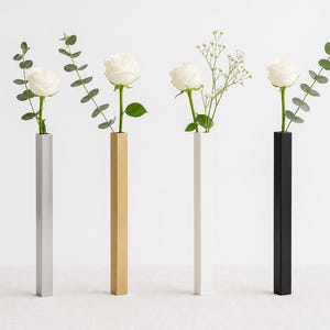 May include: Four modern, minimalist vases in silver, gold, white, and black, each holding a single white rose and greenery. The vases are tall, rectangular, and have a geometric design, perfect for home decor.