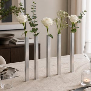 May include: Five silver metal vases, each holding a single stem of greenery or a white rose. The vases are arranged in a row on a beige tablecloth, creating a minimalist and elegant centerpiece. The scene is set for a dining experience.