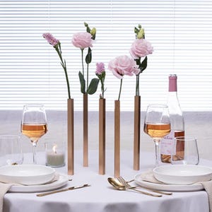 Może przedstawiać: Nakrycie stołu z miedzianymi, prostokątnymi wazonami z różowymi kwiatami. Kieliszki do wina, butelka różowego wina, białe talerze i złote sztućce ułożone na białym obrusie. Zapalona świeca.