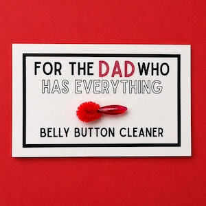 Puede incluir: Una tarjeta blanca con borde negro y texto que dice "FOR THE DAD WHO HAS EVERYTHING BELLY BUTTON CLEANER" sobre un fondo rojo. Un limpiador de ombligo rojo está en el centro de la tarjeta.