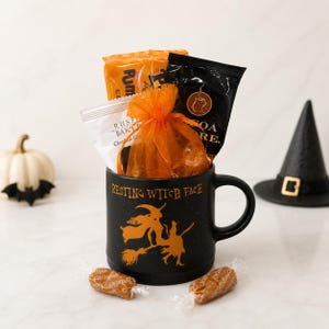 May include: A black ceramic mug featuring the text Resting Witch Face and an orange silhouette of a witch on a broomstick. The mug is filled with an orange organza bag, a black cocoa packet, and an orange snack bag. Two caramel candies lie in front.