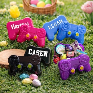 May include: A collection of colorful, game controller-shaped containers, each with a name on top. The controllers are in pink, blue, black, and purple, with yellow and black buttons. Easter eggs and a basket are in the background, set on green grass.