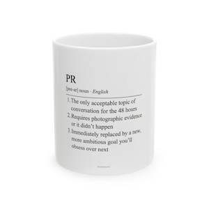 May include: A white ceramic mug featuring black printed text that defines the term PR. The text includes a phonetic pronunciation, the noun classification, and a three-point list describing the term with humorous, cynical commentary on modern work culture.