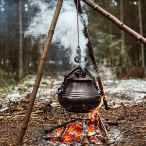 Puede incluir: Una olla de metal oscuro suspendida sobre una fogata. La olla cuelga de un trípode de ramas de madera, con vapor saliendo de ella. El fuego de abajo tiene llamas de color naranja y rojo brillante, en un entorno forestal.