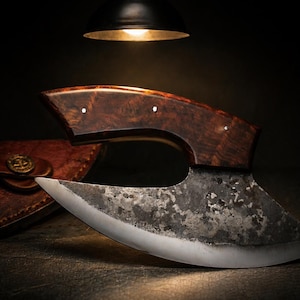 Puede incluir: Un cuchillo de carnicero artesanal con una hoja curva y un mango de madera. La hoja tiene un acabado oscuro y texturizado, y el mango presenta una veta de madera rica y oscura. Una funda de cuero es visible en el fondo.