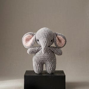 Puede incluir: Un elefante de ganchillo gris con el interior de las orejas rosa está sobre un bloque negro. El animal de peluche hecho a mano tiene pequeños ojos negros y una larga trompa. Es un juguete suave y afelpado.