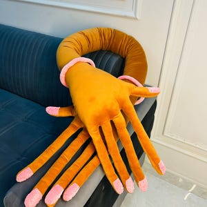May include: A plush pillow shaped like a large hand with long, extended fingers. The hand is mustard yellow with light pink fabric accents on the fingertips and a matching pink ring around the wrist area, featuring a soft, velvet-like texture.