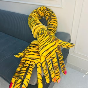 Op de afbeelding: Een pluche knuffel met lange armen en een geel-zwart tijgerstrepenpatroon. De twee grote handen hebben vingers met gekleurde stoffen nagels in rood, oranje en roze, en elke pols is versierd met een effen gele stoffen ring.