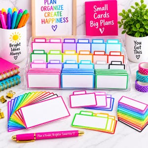 Puede incluir: Un organizador de acrílico transparente lleno de tarjetas de índice coloridas con pestañas, acompañado de un bolígrafo rosa con el texto 'Plan a Kinder, Brighter Tomorrow', una taza blanca con 'Bright Ideas Always' y una taza blanca con 'You Got This' y una planta.