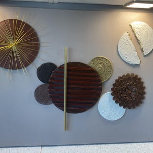 May include: A wall art piece featuring a collection of circular elements in brown, gold, and white. It includes a sunburst design with thin gold rays, a large dark brown circle with horizontal lines, a textured white crescent, and a brown floral shape.