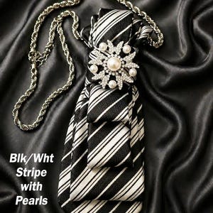May include: Black and white striped tie with a pearl and rhinestone embellishment. A silver chain is attached to the tie. The text "Blk/Wht Stripe with Pearls" is visible.