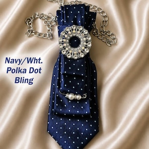 May include: Navy blue polka dot tie with a large, sparkling rhinestone brooch and a pearl pin. The tie is attached to a silver chain. The text "Navy/Wht. Polka Dot Bling" is visible in the image.