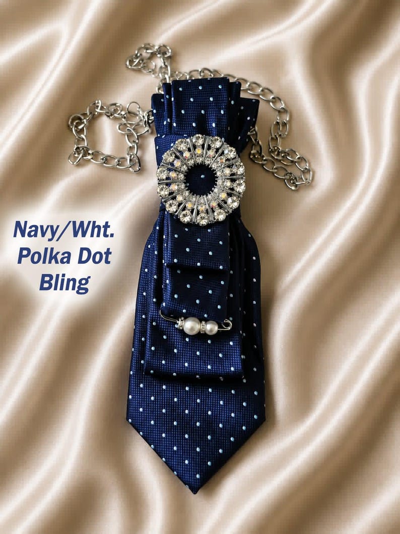 May include: Navy blue polka dot tie with a large, sparkling rhinestone brooch and a pearl pin. The tie is attached to a silver chain. The text "Navy/Wht. Polka Dot Bling" is visible in the image.