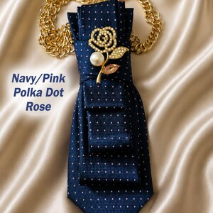 May include: Navy blue polka dot tie with a gold chain and a rose-shaped brooch. The brooch features a pearl and pink crystal accents. The text "Navy/Pink Polka Dot Rose" is visible.