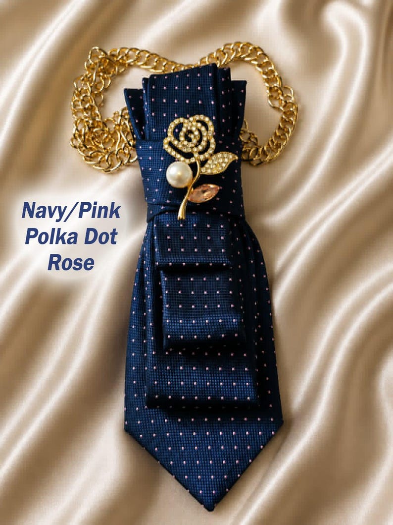 May include: Navy blue polka dot tie with a gold chain and a rose-shaped brooch. The brooch features a pearl and pink crystal accents. The text "Navy/Pink Polka Dot Rose" is visible.