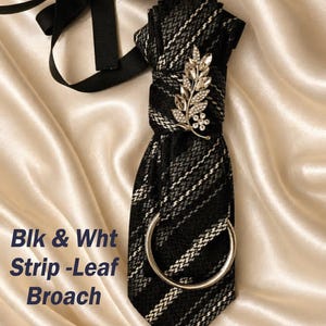 May include: A black and white striped tie with a silver leaf brooch and a silver ring. The tie has a black ribbon and the text "Blk & Wht Strip-Leaf Broach". The tie is displayed on a satin surface.