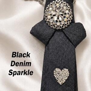 May include: Black denim tie with a silver chain necklace. The tie features a large, sparkling, round brooch and a small, heart-shaped embellishment. The text "Black Denim Sparkle" is visible.