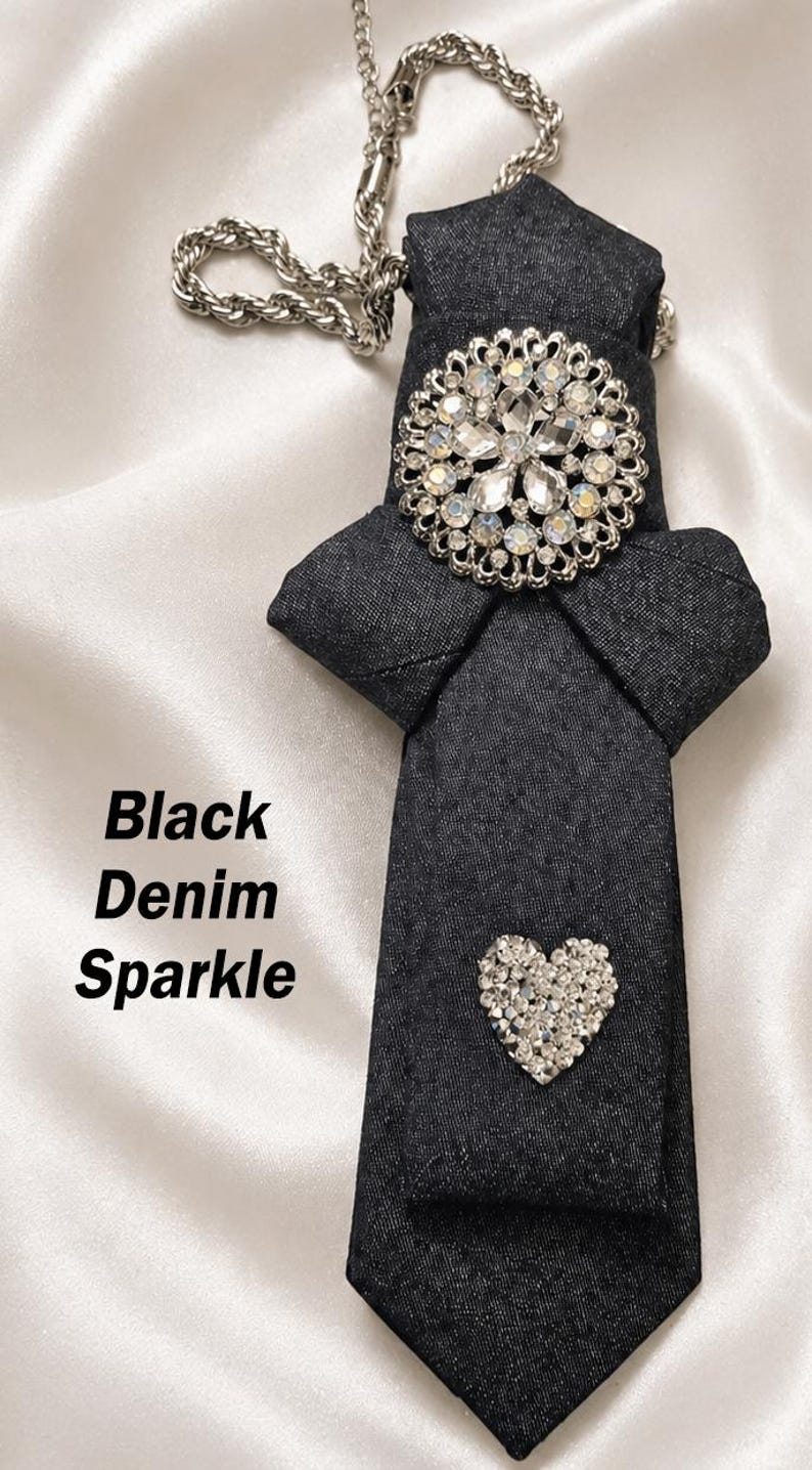 May include: Black denim tie with a silver chain necklace. The tie features a large, sparkling, round brooch and a small, heart-shaped embellishment. The text "Black Denim Sparkle" is visible.