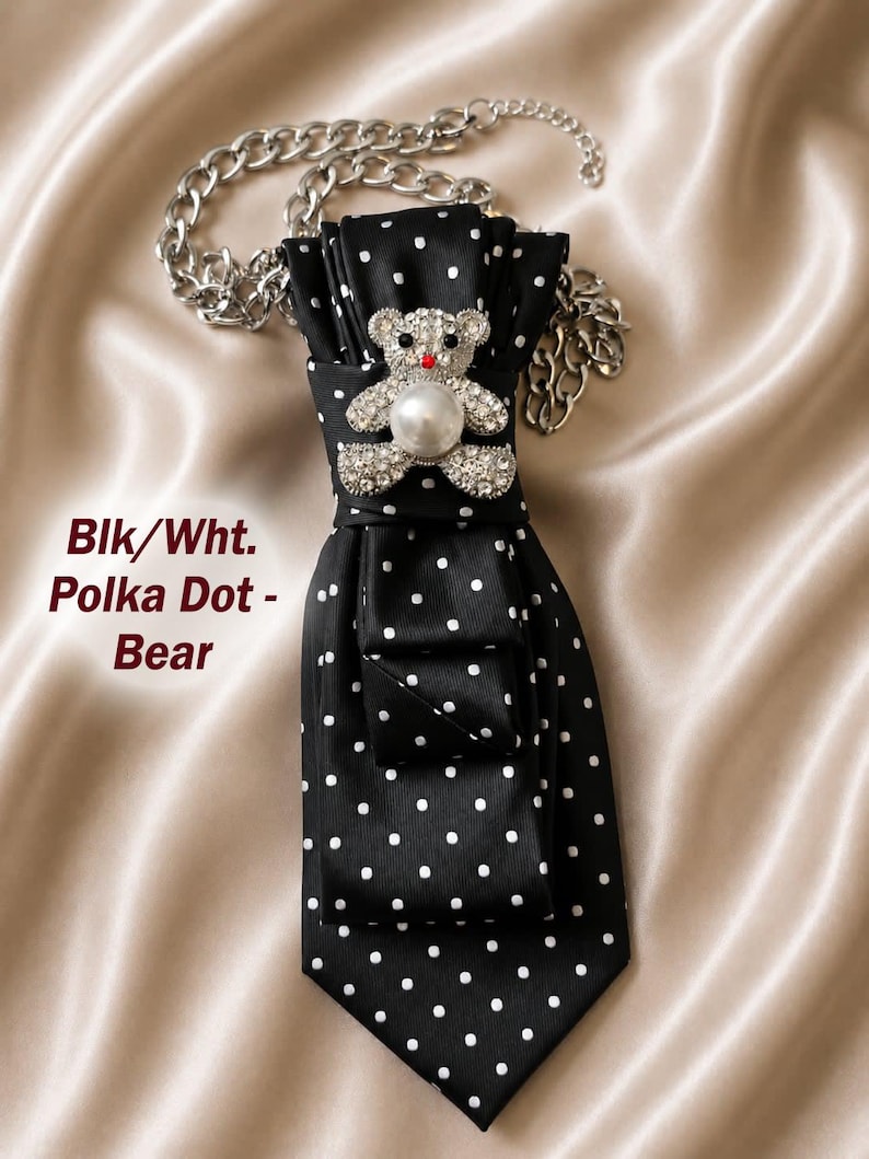 May include: A black and white polka dot tie with a silver chain necklace. The tie features a jeweled teddy bear brooch with a pearl. The text "Blk/Wht. Polka Dot - Bear" is visible.