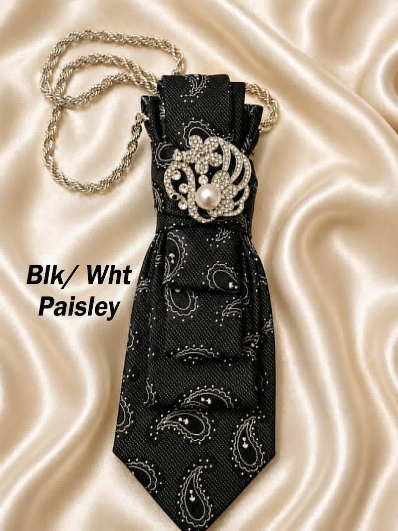 May include: Black and white paisley necktie with a decorative silver chain. The tie features a jeweled brooch with a pearl center. The text "Blk/ Wht Paisley" is visible on the left side.