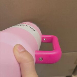 Puede incluir: Una botella de agua aislada de color rosa con un asa rosa brillante. La botella tiene una etiqueta blanca con el texto negro "WATER H2.0". El asa est&aacute; sujeta a la botella con dos remaches plateados.