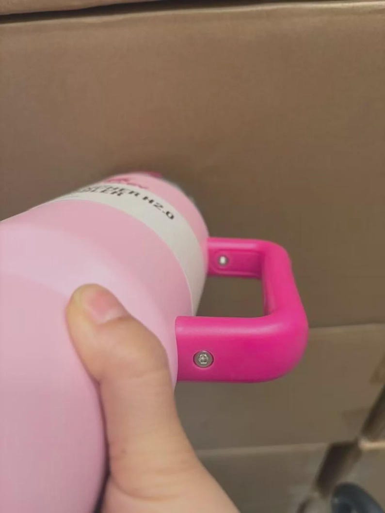 Puede incluir: Una botella de agua aislada de color rosa con un asa rosa brillante. La botella tiene una etiqueta blanca con el texto negro "WATER H2.0". El asa est&aacute; sujeta a la botella con dos remaches plateados.