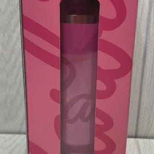 Op de afbeelding: Een roze lipstick tube wordt getoond in een roze doos met een uitgesneden venster. De lipstick heeft een verloop van roze tinten. De doos heeft een cursief ontwerp in een donkerdere roze tint.