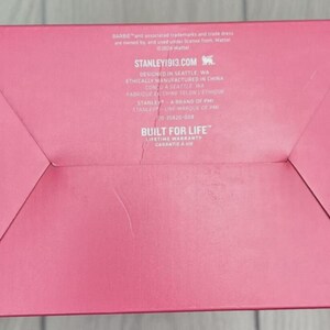 Op de afbeelding: Een roze kartonnen doos met gevouwen hoeken. De tekst "STANLEY1913.COM" is zichtbaar, samen met de zin "BUILT FOR LIFE" en een levenslange garantie. De doos heeft een matte afwerking.