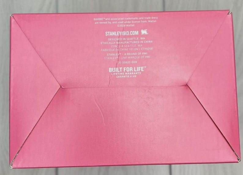 Op de afbeelding: Een roze kartonnen doos met gevouwen hoeken. De tekst "STANLEY1913.COM" is zichtbaar, samen met de zin "BUILT FOR LIFE" en een levenslange garantie. De doos heeft een matte afwerking.