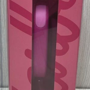 Op de afbeelding: Een roze rechthoekige doos met een helder venster dat een cilindrisch object onthult. De doos heeft een groot, gestileerd scriptontwerp in een donkerdere roze tint. Het object binnenin lijkt een schoonheidsproduct te zijn.