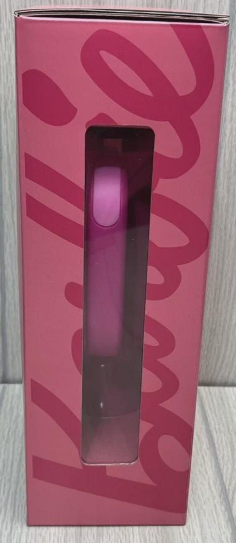 Op de afbeelding: Een roze rechthoekige doos met een helder venster dat een cilindrisch object onthult. De doos heeft een groot, gestileerd scriptontwerp in een donkerdere roze tint. Het object binnenin lijkt een schoonheidsproduct te zijn.