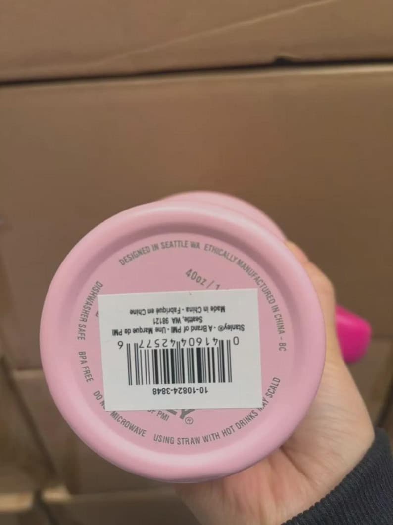 Puede incluir: Un vaso rosa de la marca Stanley, 1,18L, con una etiqueta blanca y un c&oacute;digo de barras. La parte inferior del vaso tiene el texto "Designed in Seattle WA" y "Ethically Manufactured in China".