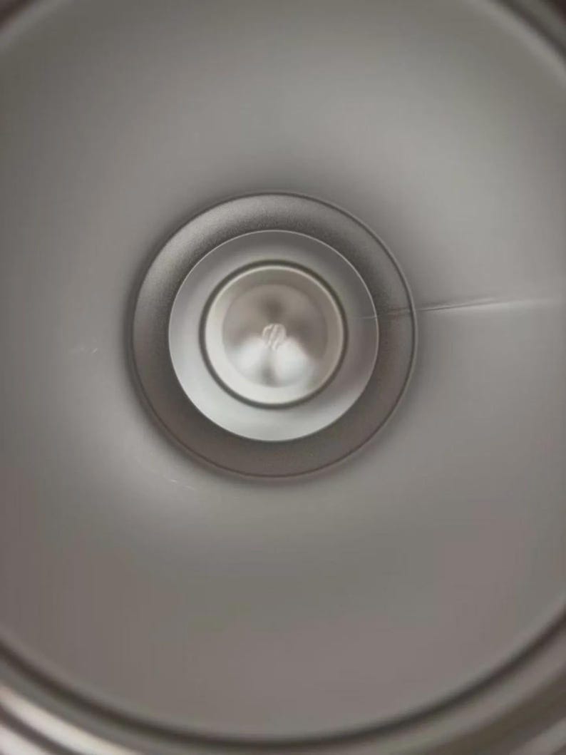 Puede incluir: Primer plano de un objeto circular plateado con anillos conc&eacute;ntricos. El centro tiene un peque&ntilde;o elemento circular reflectante. El objeto parece ser de metal y tiene una superficie lisa y pulida.