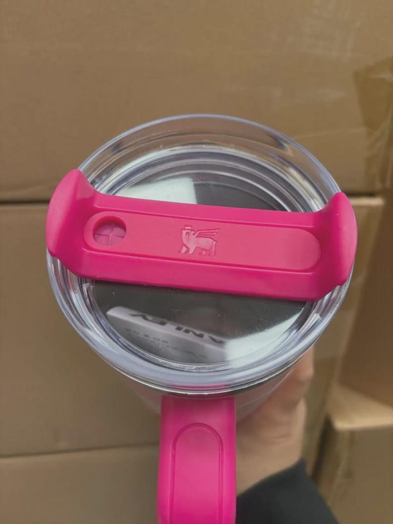 Puede incluir: Vista de cerca de un vaso transparente con tapa y asa rosas. La tapa tiene un deslizador rosa con un logotipo. El vaso est&aacute; dise&ntilde;ado para bebidas y probablemente est&aacute; aislado para mantener la temperatura.