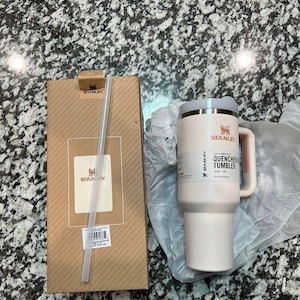 May include: A light pink Stanley Quencher tumbler with a handle and a straw, next to its cardboard packaging. The tumbler has a white lid and a pink handle. The packaging has the Stanley logo.