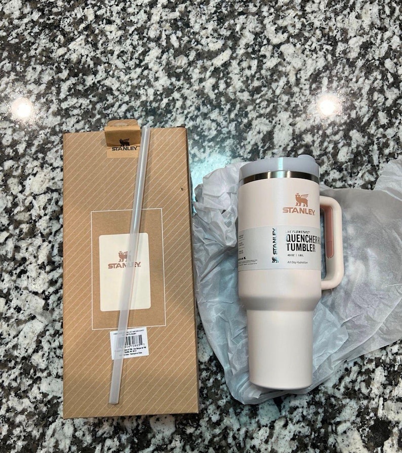 May include: A light pink Stanley Quencher tumbler with a handle and a straw, next to its cardboard packaging. The tumbler has a white lid and a pink handle. The packaging has the Stanley logo.