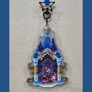 Peut inclure: Un porte-clés en acrylique transparent représentant une structure de château bleu avec des accents dorés et trois gemmes rouges circulaires à la base. Le design comprend un fermoir noir en forme d'étoile. Le château a des tours pointues.