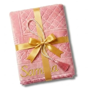 Puede incluir: Una alfombra de oración rosa con un diseño acolchado, envuelta con una cinta y un lazo dorados. Se incluye un rosario de perlas y una borla. El nombre "Samia" está bordado en hilo dorado.