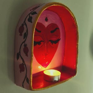Può includere: Una mensola da parete ad arco con esterno rosa chiaro caratterizzato da un'illustrazione di vite e melograni neri. L'interno è dipinto di rosso brillante con un design a forma di cuore. Una piccola candela tea light accesa poggia sulla base.