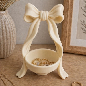 Puede incluir: Soporte para anillos de cerámica color crema con forma de lazo. El soporte tiene un cuenco pequeño para guardar anillos, con varios anillos de oro en su interior. Los lados con forma de cinta del lazo se curvan hacia abajo para formar la base.