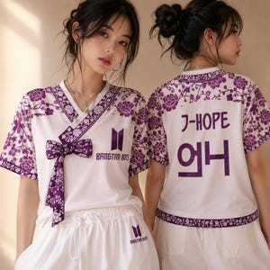 May include: A white short-sleeved top with a purple floral pattern on the collar, sleeves, and hem. The front features a bow, the text BANGTAN BOYS, and a logo. The back displays the text J-HOPE and Korean characters in purple.