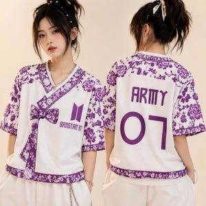 Puede incluir: Una camiseta blanca de cuello en V con patrones florales morados, con un frente estilo cruzado con un lazo, el texto BANGTAN BOYS y un logo. La parte posterior muestra el texto ARMY y el número 07 en letras moradas grandes sobre tela de malla.