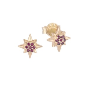 May include: A pair of gold-colored starburst stud earrings. Each earring features a central cluster of small, round, red gemstones. The earrings have a post and butterfly backing.